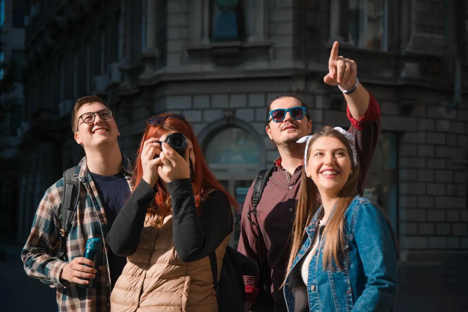 Un groupe de quatre jeunes adultes profitant d'un moment de tourisme en ville. Une personne tient un appareil photo, une autre une boisson, tandis que les autres regardent quelque chose dans le ciel. Ils semblent tous heureux, souriants et portent des lunettes de soleil, capturés dans un cadre ensoleillé avec un bâtiment historique en arrière-plan.