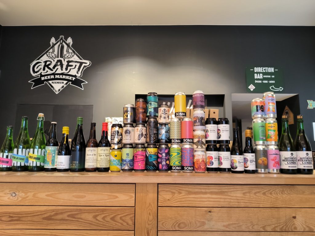 Plusieurs bouteilles et canettes de bière colorées sont alignées sur un comptoir en bois, avec un logo "Craft Beer Market Namur" en arrière-plan.