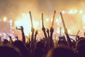 Des silhouettes de personnes brandissant des bâtons lumineux devant une scène éclairée lors d'un concert en plein air.