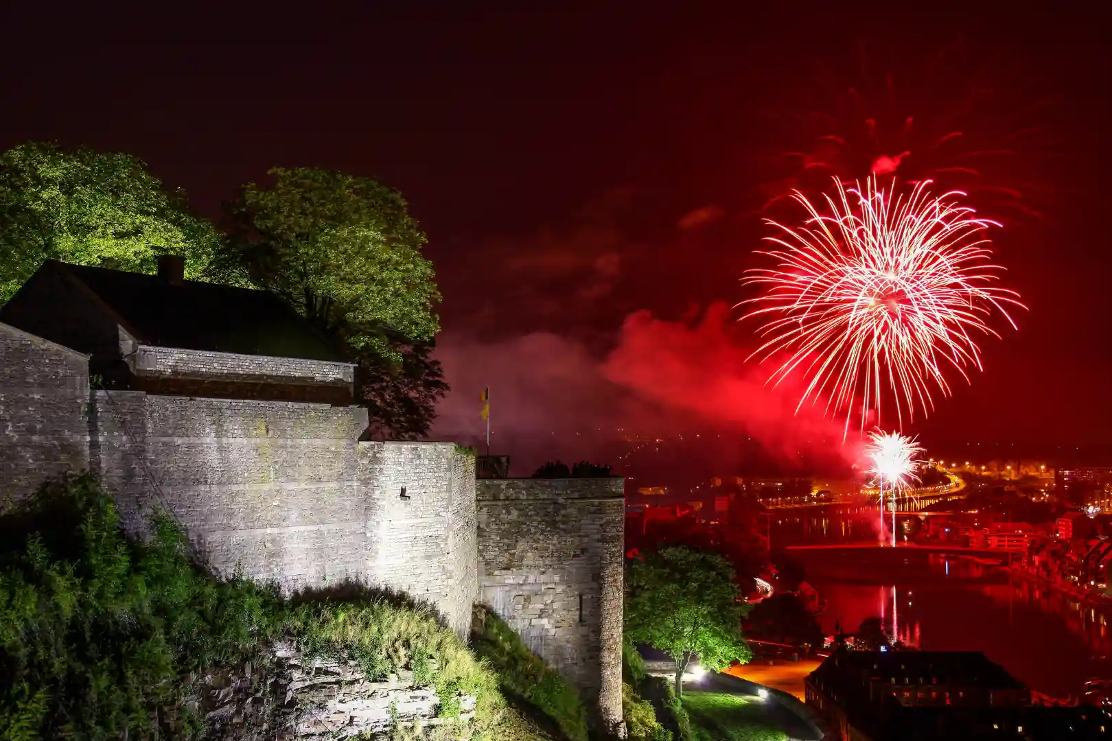 Feux d'artifice rouges illuminant le ciel nocturne au-dessus d'une forteresse en pierre éclairée, entourée de verdure, avec une rivière en arrière-plan.