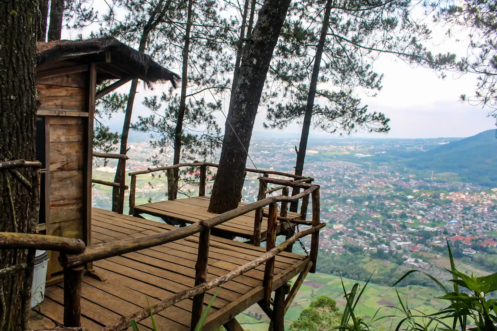 Plateforme en bois avec garde-corps entourée d'arbres, surplombant une vue panoramique d'une vallée et d'une ville en contrebas.