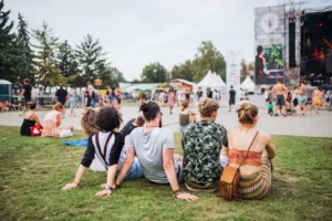 Un groupe de personnes assises sur l'herbe regarde une scène de concert en plein air avec des arbres en arrière-plan.