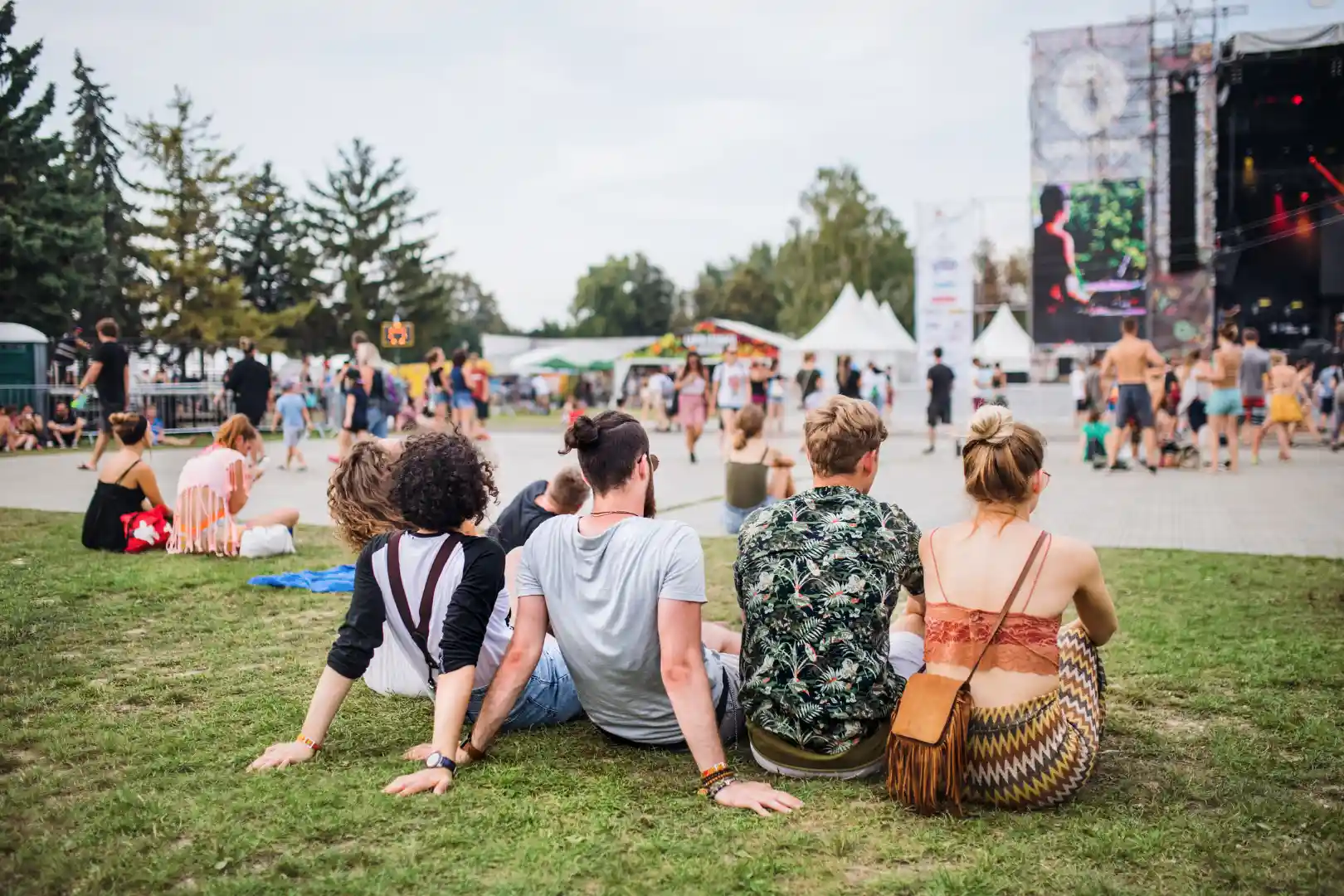 Un groupe de personnes assises sur l'herbe regarde une scène de concert en plein air avec des arbres en arrière-plan.