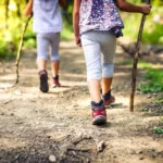 Deux enfants en pantalons gris et chaussures rouges marchent sur un sentier forestier, chacun tenant un bâton de marche.