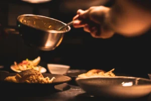 Une main verse une sauce d'une casserole en métal sur un plat de pâtes, sous un éclairage tamisé.