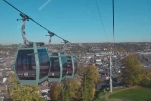 Trois cabines de téléphérique vertes survolent une ville avec des bâtiments et des arbres sous un ciel bleu clair.